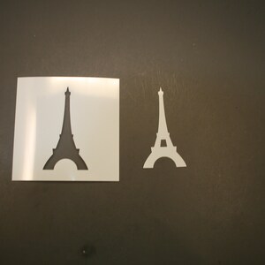 May include: Two white stencils of the Eiffel Tower on a dark gray surface. One stencil is a square with the tower cut out, and the other is a standalone cutout of the tower. The stencils are for crafting or art projects.