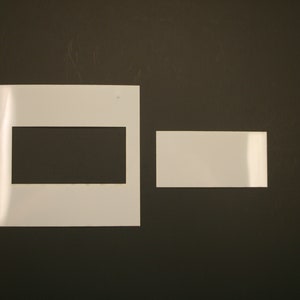 May include: Two white rectangular frames with a black rectangular opening in the larger frame. The frames are on a dark brown background.