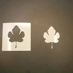 May include: Two white vinyl stencils of a leaf shape. The stencils are on a brown surface.