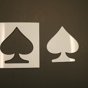 May include: Two white plastic stencils of a spade playing card symbol. The stencil on the left is on a white background, while the stencil on the right is on a brown background.