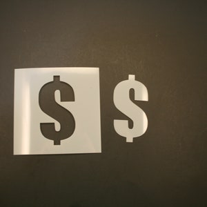 May include: Two white plastic stencils of the dollar sign, one is larger than the other. The stencils are on a dark brown surface.