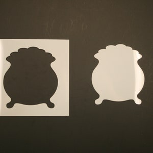 May include: Two white vinyl stencils of a cauldron, one with a solid black outline and the other with a white outline.
