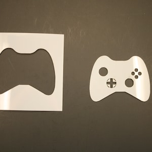 May include: Two white plastic stencils of a video game controller. The stencil on the left is larger and has a more basic design. The stencil on the right is smaller and has more detail, including buttons and a directional pad.