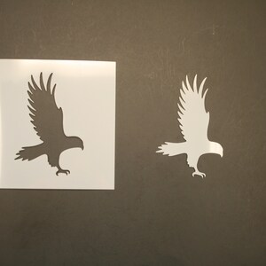 May include: An eagle stencil in two variations. One is a white eagle cut-out, and the other is a black eagle silhouette on a white square. The eagle is depicted with outstretched wings, ready for flight.