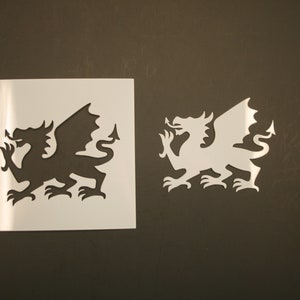 May include: Two white stencils of a Welsh dragon, one larger than the other. The stencils are on a dark brown background.
