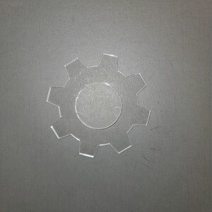 May include: A clear plastic gear with a circular hole in the center. The gear is on a gray background.