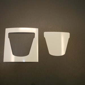 May include: Two white stencils of a flower pot on a dark gray surface. One stencil is the shape of the pot, and the other is the negative space of the pot. The stencils are for crafting and DIY projects.