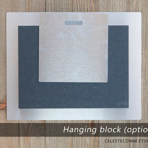 May include: A silver metal hanging block with a black foam backing. The hanging block is used to hang artwork on a wall.