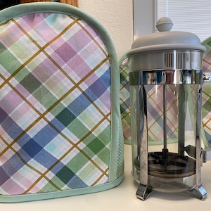May include: A gray and silver French press coffee maker with a white handle. The coffee maker is sitting on a table with a plaid fabric cover.