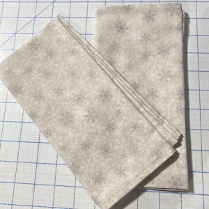 May include: Two white fabric napkins with a snowflake pattern. The napkins are folded and stacked on top of each other.