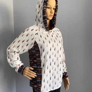 Op de afbeelding: Witte hoodie met lange mouwen. De voorkant van de hoodie heeft een patroon van vredestekens in bruin en oranje. De achterkant van de hoodie heeft een patroon van woorden in bruin, oranje en wit. De woorden omvatten "power", "equality", "freedom" en "justice".