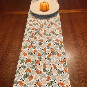 May include: A fall-themed table runner with a repeating pattern of leaves in shades of orange, green, and beige on a white background. A small orange pumpkin sits on a wooden plate in the center of the runner. The runner is placed on a dark brown wooden table.