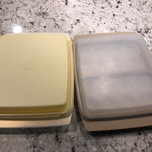 May include: Two rectangular plastic food storage containers with lids. The container on the left is yellow and the container on the right is beige.