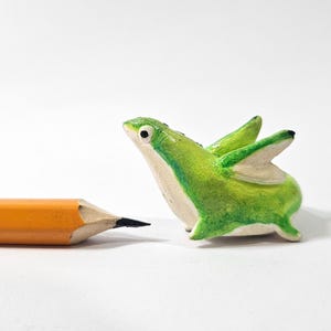 May include: A small, lime green creature with white details and wing-like features is positioned beside a yellow pencil. The creature has a whimsical design, with a textured surface and black details. The pencil is sharpened.