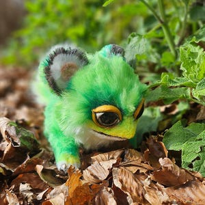 May include: A green plush creature with yellow eyes and a white belly sits amongst fallen leaves and green foliage. The creature has fluffy fur and detailed features, resembling a fantastical animal. The background is blurred, highlighting the plush toy.