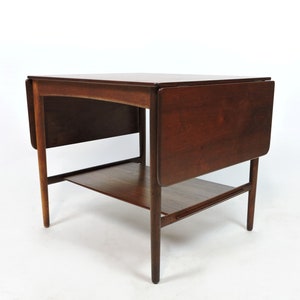 May include: A dark brown wooden drop leaf table with a lower shelf. The table has a simple, mid-century modern design.