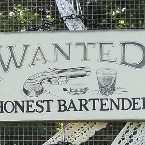 May include: Rectangular wooden sign with the words "WANTED HONEST BARTENDER" in black lettering. The sign features an illustration of a pistol, a shot glass, and a few coins. The sign is cream-colored with decorative flourishes.