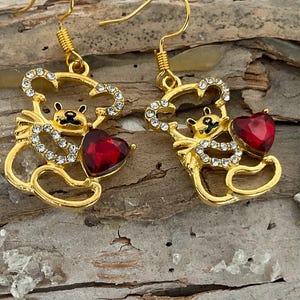 May include: Gold-tone bear earrings with red heart accents. The bear-shaped earrings are embellished with clear crystals and feature a red heart. The earrings are displayed on a piece of wood.