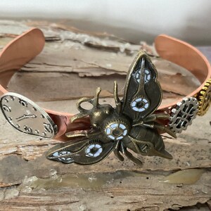 May include: A copper cuff bracelet with a steampunk-inspired design featuring a brass butterfly with white accents, gears, and a clock face.