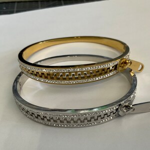 Peut inclure: Deux bracelets joncs, l'un doré et l'autre argenté, chacun avec un motif de fermeture éclair. Les fermetures éclair sont ornées de petits cristaux transparents. Les bracelets sont présentés sur une surface blanche.