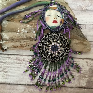 May include: A purple and green beaded necklace featuring a face pendant and a round metal charm. The face pendant has closed eyes and red lips. The charm has a floral design and a sparkly centre.