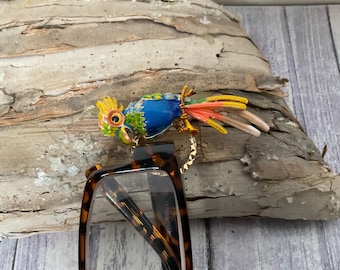 May include: A colorful parrot-shaped eyeglass holder. The parrot has a yellow crest, blue body, and multi-colored tail feathers. The holder is attached to a pair of tortoiseshell eyeglasses. The parrot is resting on a piece of wood.