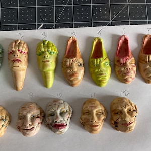 May include: Twelve small ceramic sculptures of faces with various expressions and colors. The sculptures are arranged in two rows on a white surface.