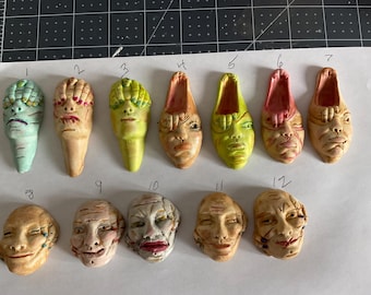 May include: Twelve small ceramic sculptures of faces with various expressions and colors. The sculptures are arranged in two rows on a white surface.
