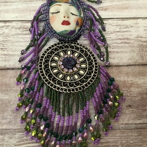 May include: A purple and green beaded necklace with a round metal pendant featuring a white centre and a purple gemstone. The pendant is attached to a ceramic face with closed eyes and red lips.