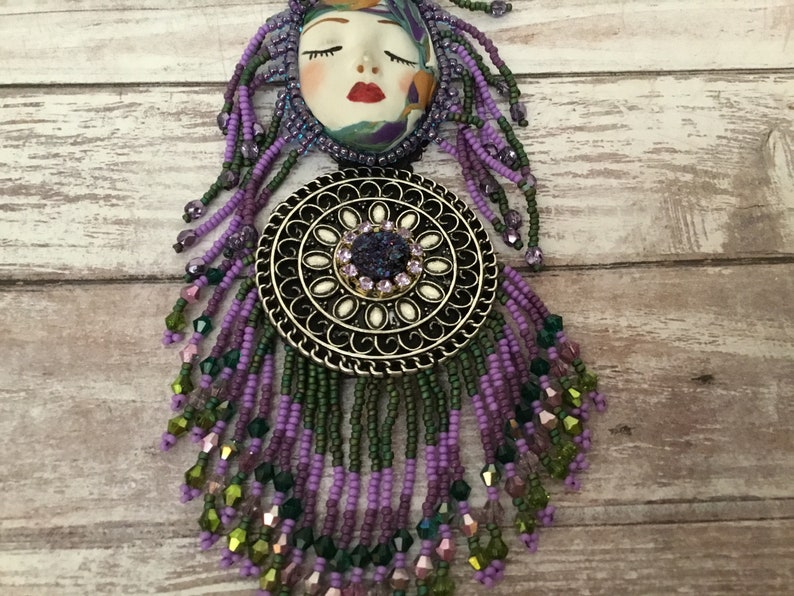 May include: A purple and green beaded necklace with a round metal pendant featuring a white centre and a purple gemstone. The pendant is attached to a ceramic face with closed eyes and red lips.