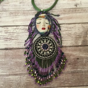 May include: A purple and green beaded necklace with a silver pendant featuring a face and a circle of rhinestones.
