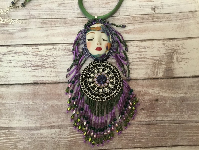 May include: A purple and green beaded necklace with a silver pendant featuring a face and a circle of rhinestones.