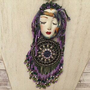 May include: A purple and green beaded necklace featuring a ceramic face pendant and a round metal charm. The face has closed eyes and red lips.