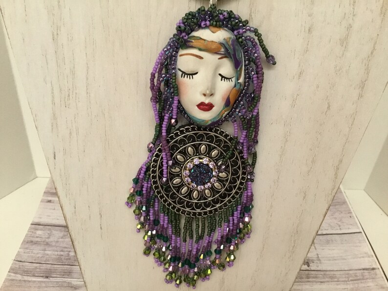 May include: A purple and green beaded necklace featuring a ceramic face pendant and a round metal charm. The face has closed eyes and red lips.