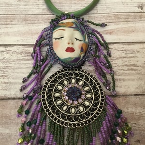 May include: A purple and green beaded necklace with a silver pendant featuring a white circle with a purple rhinestone in the centre. The pendant is attached to a ceramic face with closed eyes and red lips.