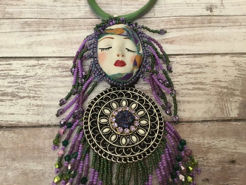 May include: A purple and green beaded necklace with a silver pendant featuring a white circle with a purple rhinestone in the centre. The pendant is attached to a ceramic face with closed eyes and red lips.