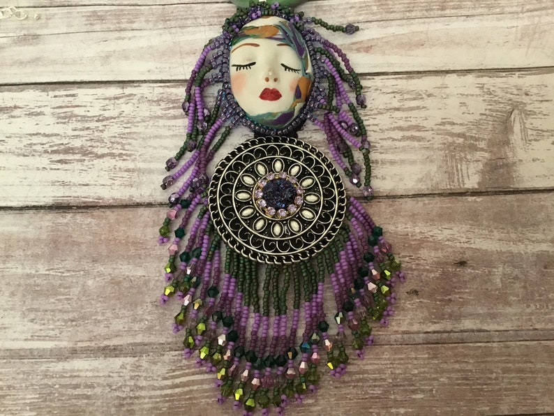 May include: A purple and green beaded necklace featuring a face charm and a round metal pendant. The pendant has a floral design and a purple gemstone in the centre.