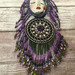 May include: A purple and green beaded necklace with a silver pendant featuring a face and a circular design. The pendant has a large, sparkly purple stone in the centre.