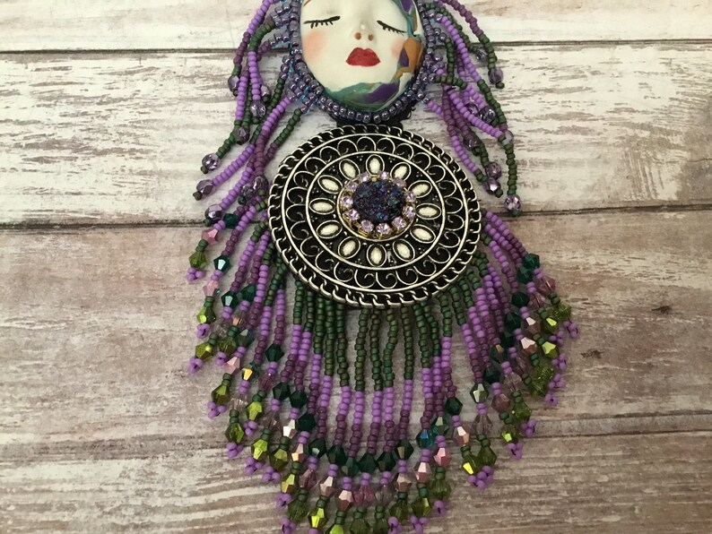 May include: A purple and green beaded necklace with a silver pendant featuring a face and a circular design. The pendant has a large, sparkly purple stone in the centre.