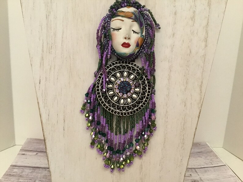 May include: A purple and green beaded necklace with a silver pendant featuring a face with closed eyes and a floral design. The pendant is surrounded by a circle of smaller beads.