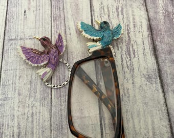 May include: Decorative hummingbird eyeglass holders. One hummingbird is purple and white, the other is blue and white. The birds are attached to a silver ring that slides onto the glasses. The glasses have a tortoise shell pattern.