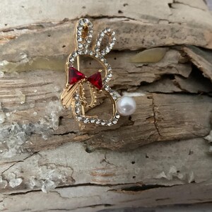 May include: A gold-toned brooch shaped like a rabbit with a red bow and a pearl accent.