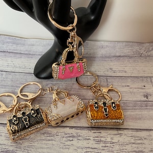 May include: Four gold-tone handbag keychains with rhinestones. The handbags are pink, white, black and brown with a leopard print, and orange.