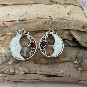 May include: A pair of silver earrings with a crescent moon design and a red gemstone accent. The moon is white and has a face sculpted on it.