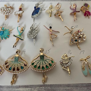 May include: A collection of eighteen gold-tone metal brooches shaped like ballerinas and ballet slippers, each numbered from 1 to 18. The designs feature various colored enamel, clear rhinestones, and faux pearls, depicting dancers in different poses.