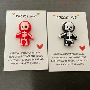 May include: Two small skeleton figurines, one red and one black, sit atop cards that read "Pocket Hug." The cards also include the text "Here's a little pocket hug, please keep it safe and close. Then it will be there beside you when you need it most."