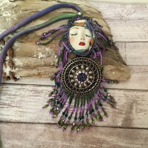 May include: A beaded necklace with a purple and green colour scheme. The pendant features a face with closed eyes and a red mouth. The pendant is surrounded by a silver-toned circular design with a large crystal in the centre.
