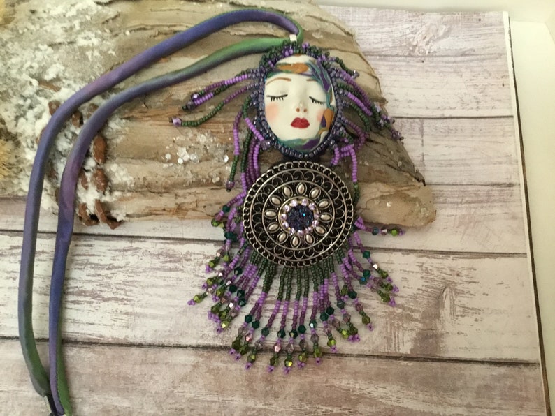 May include: A beaded necklace with a purple and green colour scheme. The pendant features a face with closed eyes and a red mouth. The pendant is surrounded by a silver-toned circular design with a large crystal in the centre.