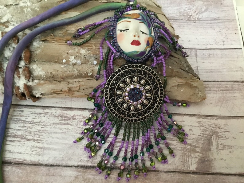May include: A purple and green beaded necklace featuring a face pendant and a round metal charm. The face pendant has closed eyes and red lips. The charm has a floral design and a sparkly centre.