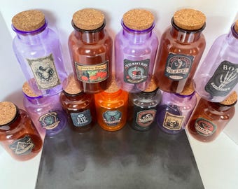 May include: A collection of eleven glass apothecary jars in various colors including purple, orange, and amber, each topped with a cork stopper. The jars feature decorative labels with text such as Pumpkin Syrup, Dead Man's Blood, Eye of Newt, and Bone Powder.